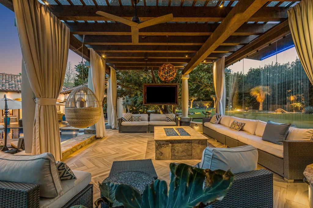 The covered outdoor lounge at dusk: a timber pergola with a hanging rattan chair, a large sectional, a stone fire and coffee table, and the rain-curtain feature and pool at left.