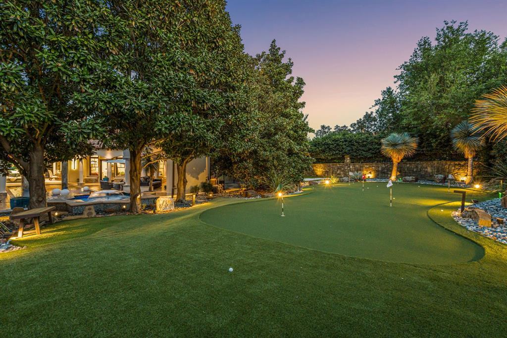 The backyard putting green at dusk: a professional-grade multi-hole green with flags, a lit stone retaining wall, magnolias and palms, and the house glowing at left.