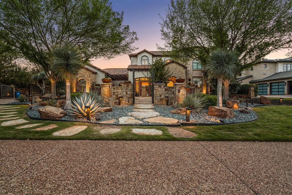 Front elevation at dusk: a stone-and-stucco two-story façade, an iron-and-glass entry, sculpted agave and yucca xeriscape, and an uplit flagstone walk.