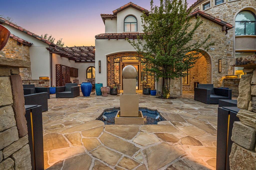 The entry courtyard at dusk: flagstone paving, a carved stone sphere fountain, an iron-and-glass front door glowing warm beneath an arched stone loggia.