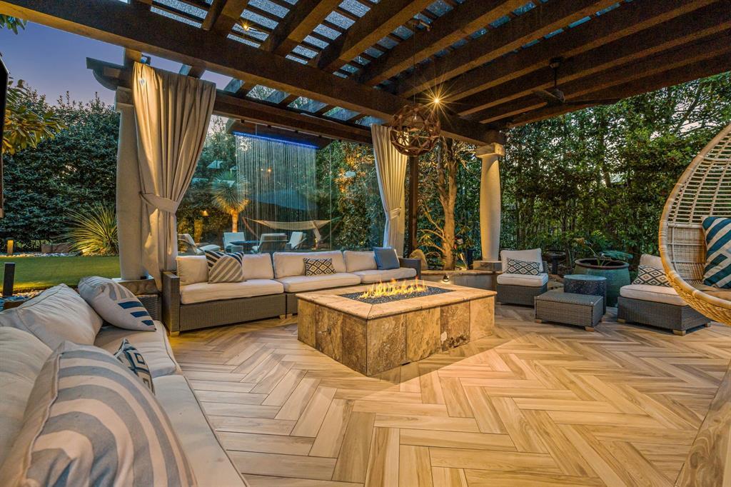 The outdoor lounge with a lit fire table: a herringbone wood-look deck, wicker sectionals, a square gas fire pit ablaze, a pergola, a hanging egg chair, and a green lawn beyond.