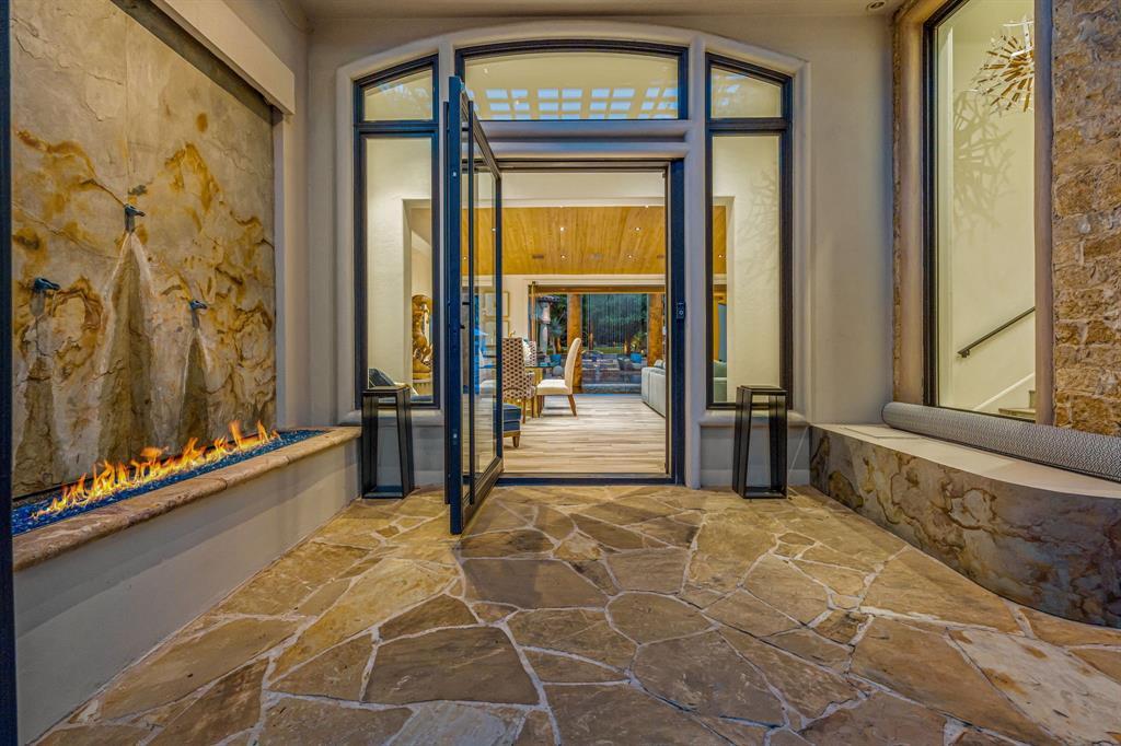The entry vestibule looking in: a linear fire-on-glass fireplace set into a raw stone slab wall, flagstone floor, and an iron-and-glass door open to the great room and pool beyond.