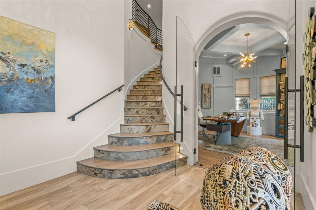 The curved staircase with book-matched stone risers and a glass rail, viewed from the hall, with the office visible through a glass wall beyond.