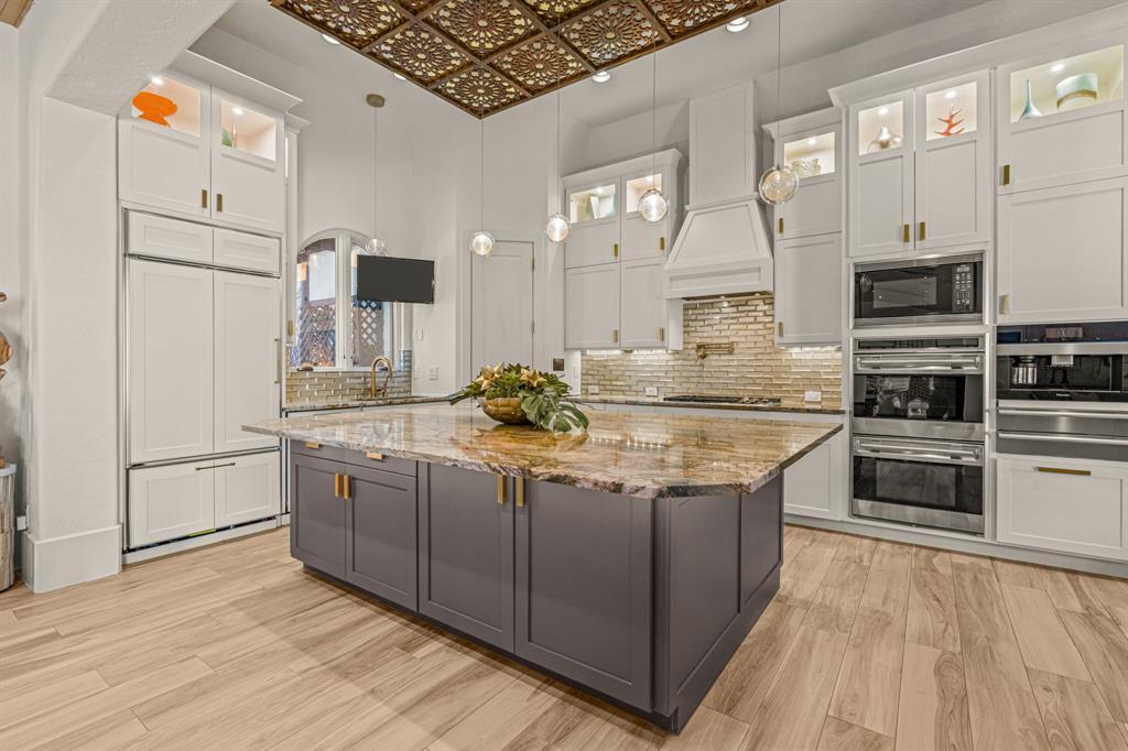 The kitchen from the side: the gray island and gold ceiling, a panel-front refrigerator, the double wall ovens and built-in coffee system, and lit glass upper cabinets.