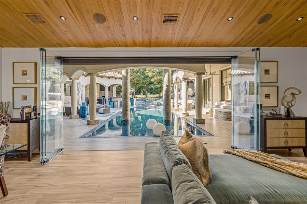 A living space with a pale wood-plank ceiling and a folding glass wall fully retracted onto the courtyard pool and stone columns, a green velvet sofa in the foreground.