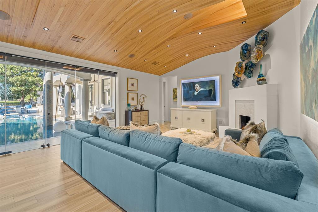 The living room: a sculptural curved wood-plank ceiling with cove lighting, a teal sectional, a backlit television over a white fireplace, and a wall of glass sliding open to the pool.