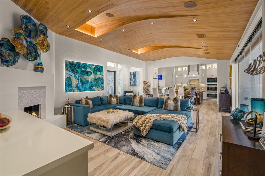 The family room: a sweeping wood-plank wave ceiling with cove lighting, a teal sectional, a white fireplace, and a wall of glass sliding open to the pool.