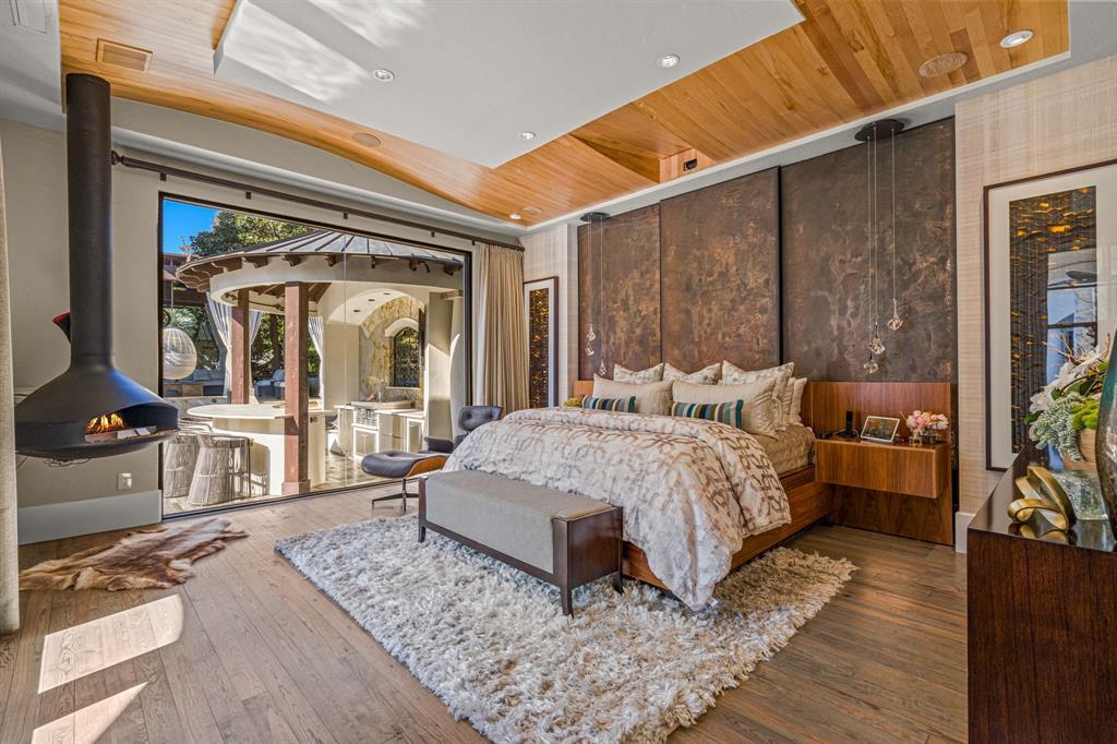 The primary suite: a patinated-copper panel headboard wall, floating walnut nightstands with suspended pendant lights, a black suspended gas fireplace, and a sliding glass wall open to the pool and cabana.