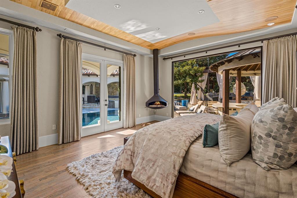 The primary bedroom toward the glass: a freestanding black cone fireplace, curtained French doors and a sliding wall opening onto the pool and cabana at dusk.