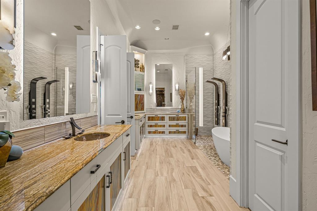 The spa bath wide: a long stone-topped dual vanity, wave-textured white stone walls, matte-black wall-mount fixtures, and a freestanding sculptural soaking tub on a pebble floor.