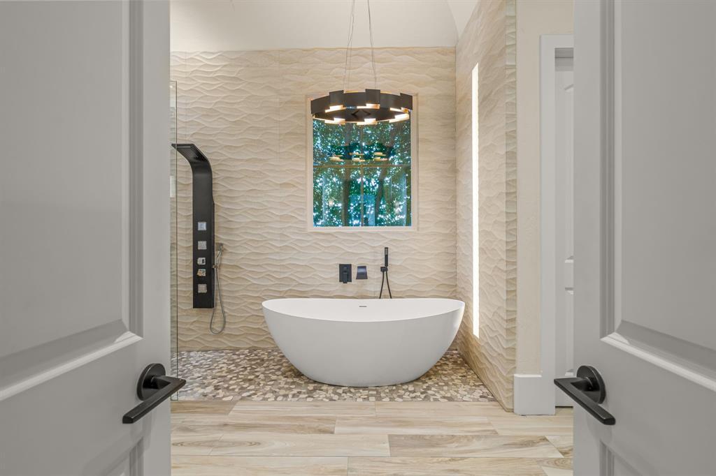The spa primary bath: a white sculptural freestanding soaking tub beneath an iron ring chandelier and an arched window, wave-textured white stone walls, and a pebble-tile floor.