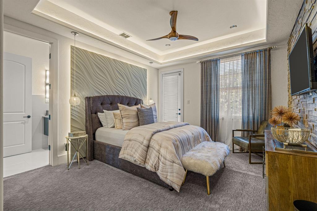 A secondary bedroom: a sculpted wavy plaster accent wall behind a tufted upholstered bed, a tray ceiling with cove light, and an en-suite door.