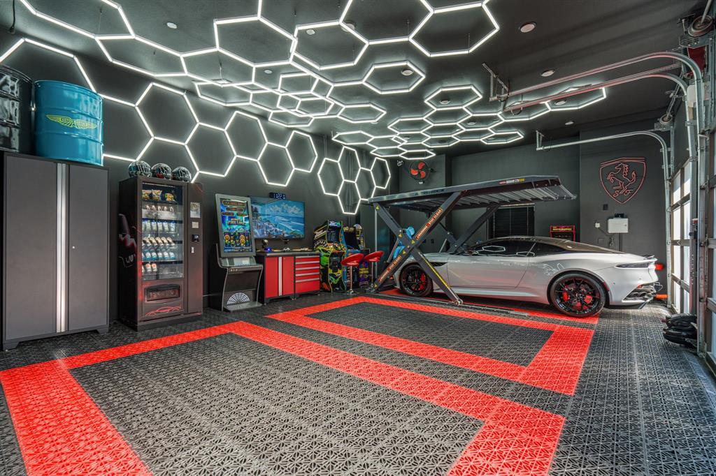 The finished three-car show garage: a hexagonal LED ceiling grid, patterned floor tiles, a scissor car lift with a silver sports car, vintage arcade cabinets, and storage cabinets.