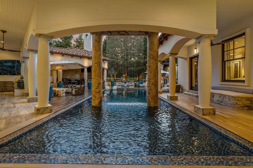 The covered pool passage: pool water flowing beneath the stone-columned loggia with a rain-curtain water feature, arches framing the outdoor lounge and kitchen beyond.