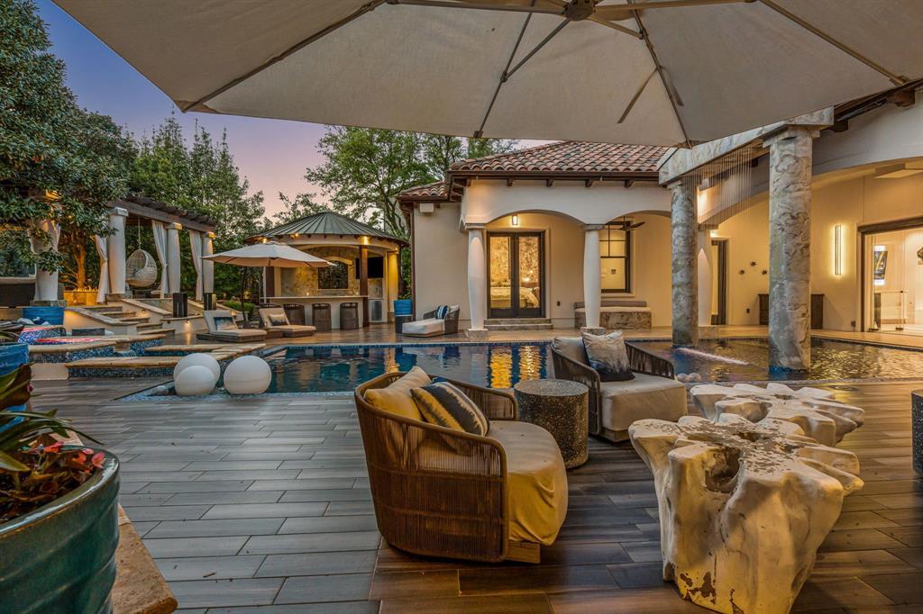 Poolside from under a cantilever umbrella at dusk: the pool, the raised spa, a covered bar and kitchen cabana with a grille, loungers, and the arched loggia.