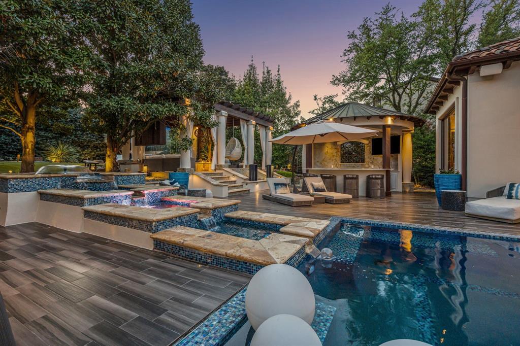The pool, spa, and fire terrace at dusk: a multi-level travertine deck, a tiled spa with LED fire-color water, a stepping-stone spillway, umbrellas, and a bar cabana under a peaked roof.