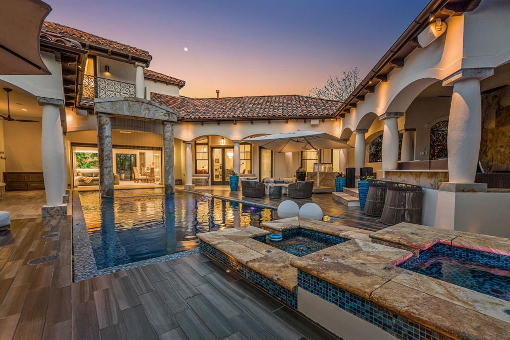The courtyard pool and raised spa at dusk: the pool in the foreground with a tiled spa and fire bowls, an arched loggia and balcony above, and warm interior glow.