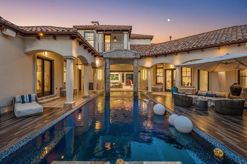 The courtyard pool centered at dusk: a long reflecting pool running straight to the stone-columned loggia and lit great room beyond, floating orbs on the water, twin roof wings mirrored.