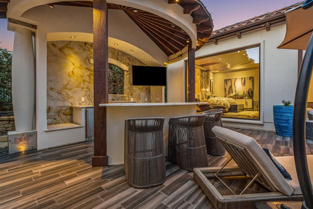 An outdoor kitchen and bar cabana: a peaked wood-slat ceiling, a backlit onyx-stone bar wall, wicker barstools, a built-in grille, and a picture window into the primary bedroom.