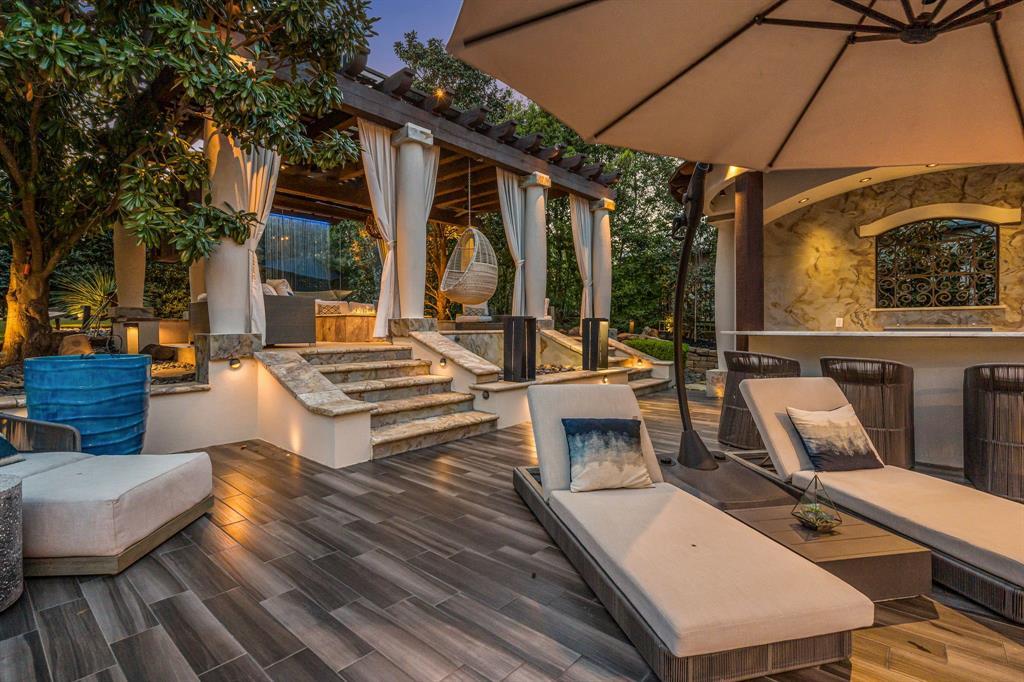 The poolside cabana lounge at dusk: a teak-tone deck, chaise loungers, a pergola with curtains and a hanging egg chair, and stone steps up to a bar with an iron-grille niche.